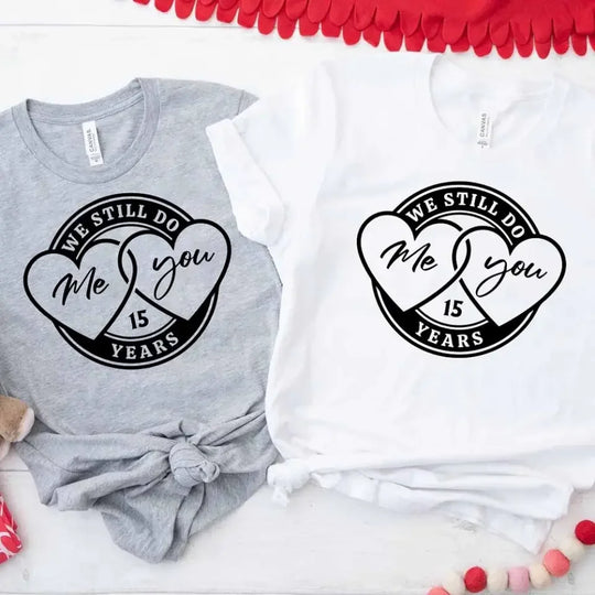 Two t-shirts, one gray and one white, each with a heart and the text 'We Still Do Me & You', displayed on a surface with decorative items around them.
