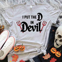 Gray t-shirt with 'I Put the D in Devil' text and Halloween-themed decorations on a textured surface.