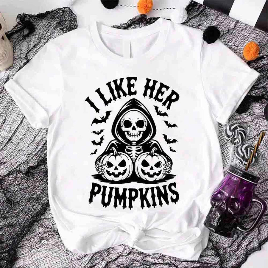 White t-shirt with Halloween-themed design featuring a skeleton holding pumpkins and text 'I Like Her Pumpkins' on a spider web background.