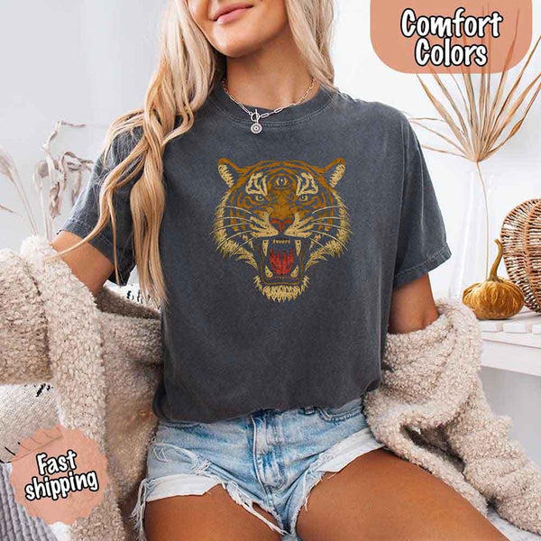 May include a woman wearing a pepper Comfort Colors T-shirt featuring a bold tiger graphic with 90s jungle safari vibes, styled casually with denim for a retro outdoor look.