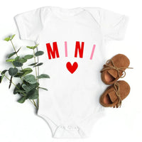 White baby onesie with 'MINI' and a heart, paired with brown shoes and green leaves on a white background