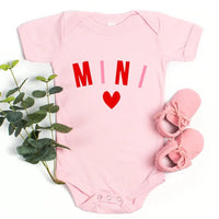 Pink baby onesie with 'MINI' and a heart, accompanied by pink shoes and a plant on a white background