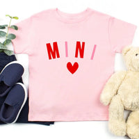 Pink t-shirt with 'MINI' and a heart, surrounded by navy shoes and a beige teddy bear on a white background