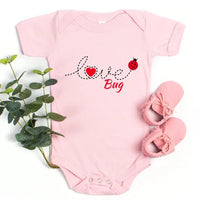 Pink baby onesie with 'Love Bug' text and ladybug design, accompanied by pink baby shoes and a plant on a white background.