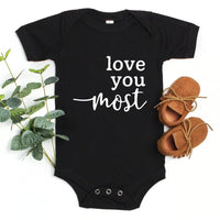 Black baby onesie with 'love you most' text, placed on a white background with shoes and plant for scale.