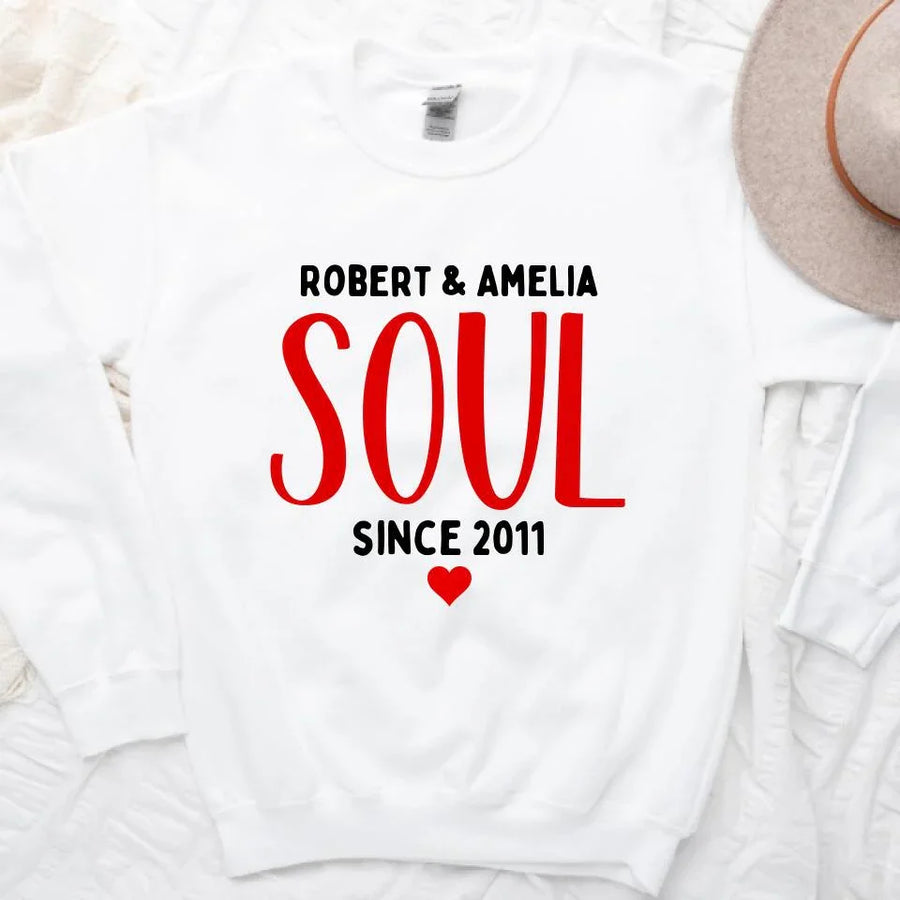 A white couple's t-shirt with the text 'ROBERT & AMELIA SOUL SINCE 2011' printed on the front, displayed on a table with denim jeans, a camera, and a plant in the background.