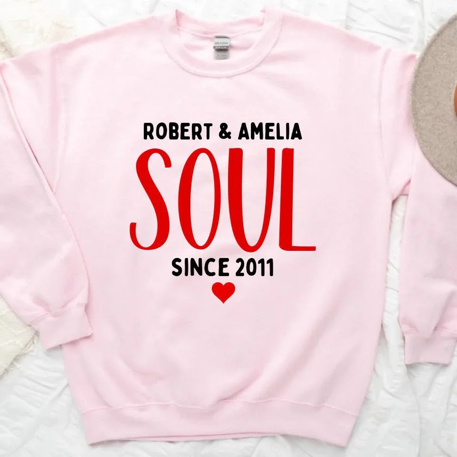 A white couple's t-shirt with the text 'ROBERT & AMELIA SOUL SINCE 2011' printed on the front, displayed on a table with denim jeans, a camera, and a plant in the background.