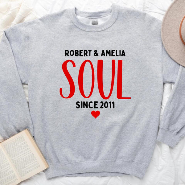 A white couple's t-shirt with the text 'ROBERT & AMELIA SOUL SINCE 2011' printed on the front, displayed on a table with denim jeans, a camera, and a plant in the background.