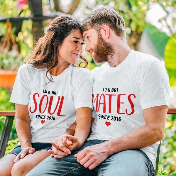 A white couple's t-shirt with the text 'ROBERT & AMELIA SOUL SINCE 2011' printed on the front, displayed on a table with denim jeans, a camera, and a plant in the background.