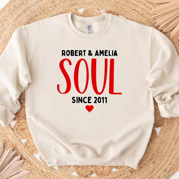 A white couple's t-shirt with the text 'ROBERT & AMELIA SOUL SINCE 2011' printed on the front, displayed on a table with denim jeans, a camera, and a plant in the background.