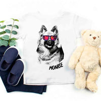 White pillow with a dog illustration and 'MONIE' text, surrounded by navy slippers, a plant, and a teddy bear on a white background.