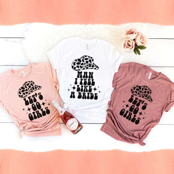 Three t-shirts in different colors with a graphic text 'Man I feel like a BRIDE' printed on them, displayed on a white surface with flowers and a watch.