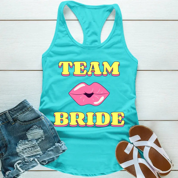 A sky blue tank top with 'TEAM BRIDE' text and a heart symbol replacing the word 'TEAM'. The tank top is displayed alongside denim shorts and white sandals, suggesting a casual outfit for a bachelorette party.