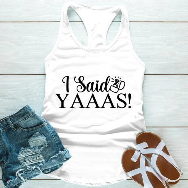 A white tank top with the text 'I Said YAAAS!' printed in black, placed on a wooden surface with blue denim shorts and white sandals beside it.