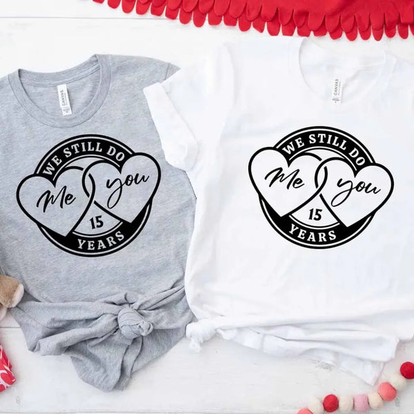 Two t-shirts, one gray and one white, each with a heart and the text 'We Still Do Me & You', displayed on a surface with decorative items around them.