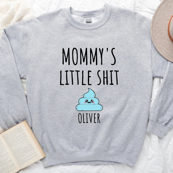 A white t-shirt with the text 'MOMMY'S LITTLE SHIT' printed on the front, above a cartoon graphic of a cloud with the name 'Oliver' added below the graphic.