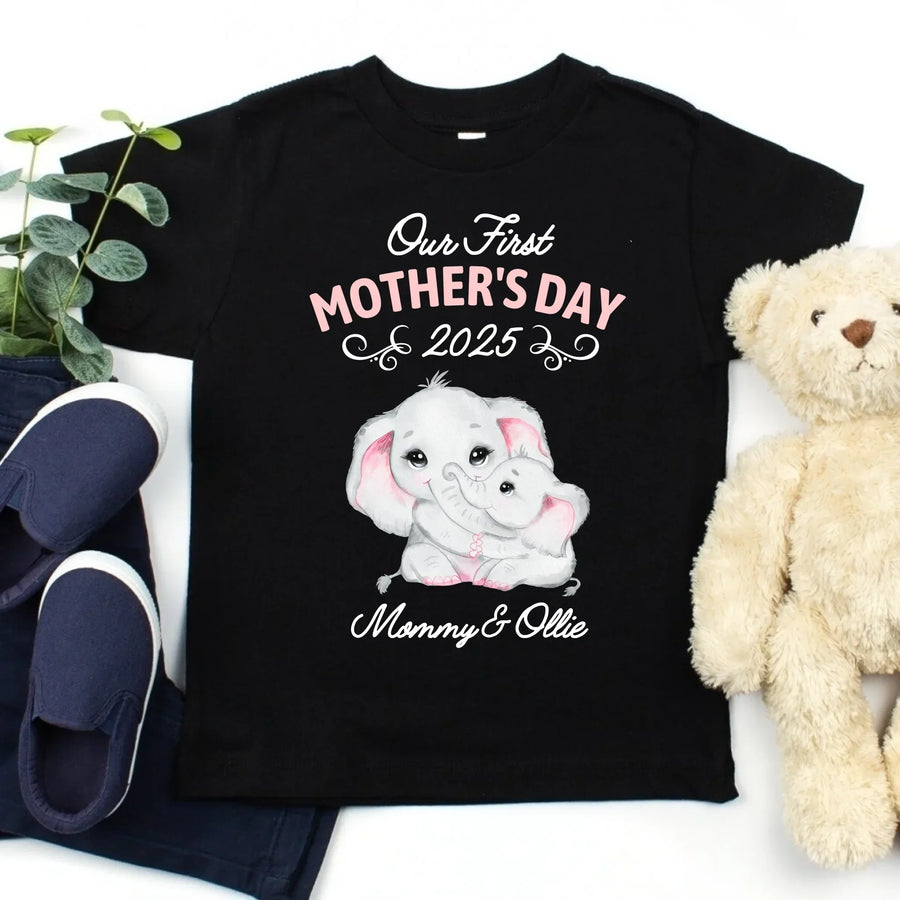 Black t-shirt with 'Our First Mother's Day 2025 Mommy & Ollie' design on a white background with shoes and a teddy bear.