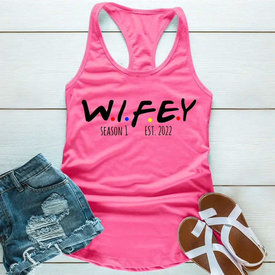 Pink tank top with 'WIFEY' text, denim shorts, and sandals on a wooden surface