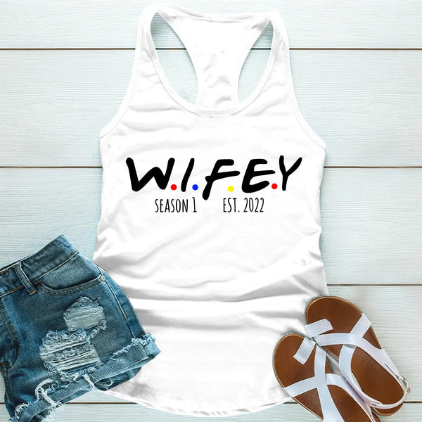 White tank top with 'WIFEY' text, denim shorts, and sandals on a light wooden surface