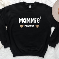 A black t-shirt with the text 'MOMMIE · roberto' printed on the front, displayed on a flat lay with denim jeans, a camera, and a plant in the background.