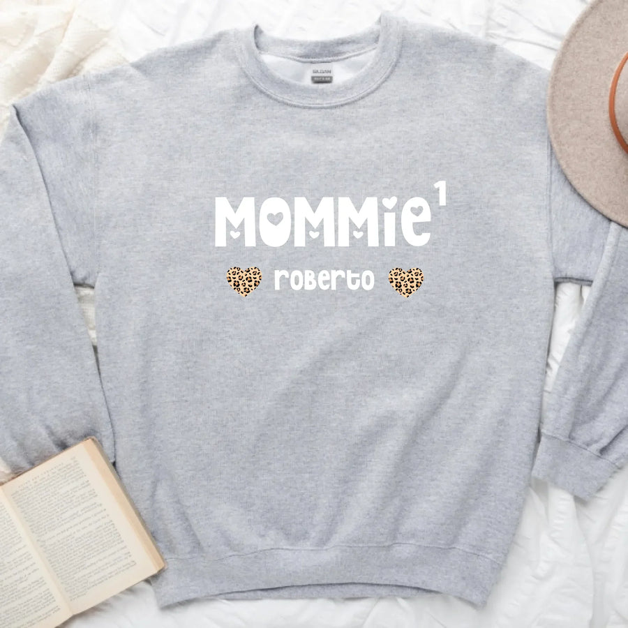 A black t-shirt with the text 'MOMMIE · roberto' printed on the front, displayed on a flat lay with denim jeans, a camera, and a plant in the background.