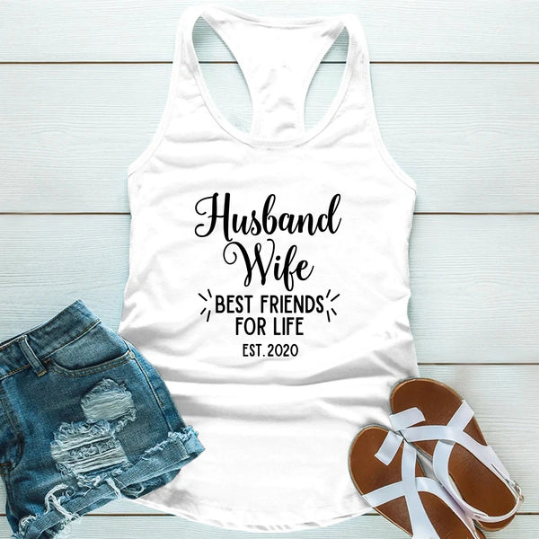 White tank top with 'Husband Wife Best Friends for Life Est. 2020' text, paired with denim shorts and sandals on a light wooden surface.