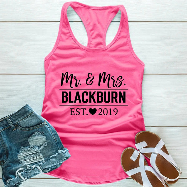 Pink tank top with 'Mr. & Mrs. Blackburn Est. 2019' text, denim shorts, and sandals on a wooden surface.