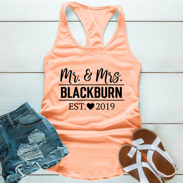 Peach tank top with 'Mr. & Mrs. Blackburn EST. 2019' text, denim shorts, and sandals on a light wooden surface.