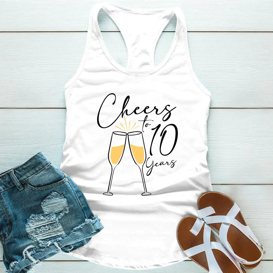White tank top with 'Cheers to 10 Years' text and champagne glass design, paired with denim shorts and sandals on a light wooden surface.