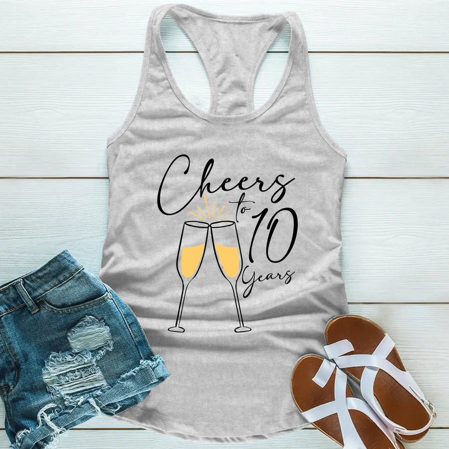 Gray tank top with 'Cheers to 10 Years' text and champagne glasses, paired with denim shorts and sandals on a light wooden surface.