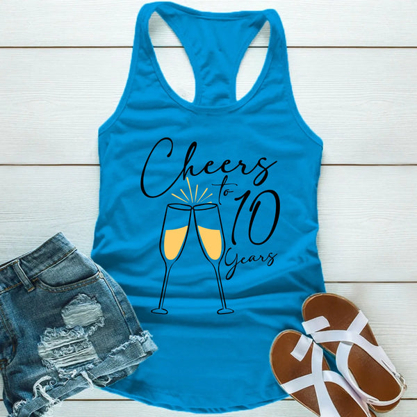 Blue tank top with 'Cheers to 10 Years' text and champagne glasses, paired with denim shorts and sandals on a wooden surface.