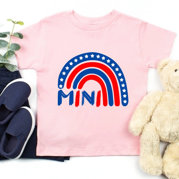 Pink t-shirt with rainbow and 'MINI' design, surrounded by navy shoes and a beige teddy bear on a white background.