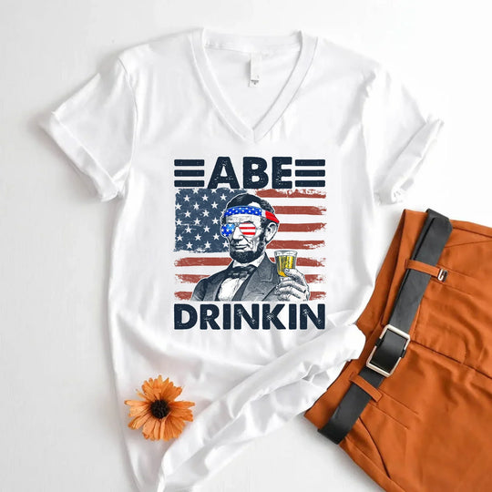 White graphic tee with Abraham Lincoln holding a drink, USA flag background, text Abe Drinkin.