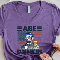 Purple tee featuring Abraham Lincoln with patriotic sunglasses, beer, and USA flag graphic