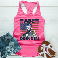 Pink tank top with Abraham Lincoln graphic wearing American flag sunglasses, holding a drink, paired with denim shorts and sandals.