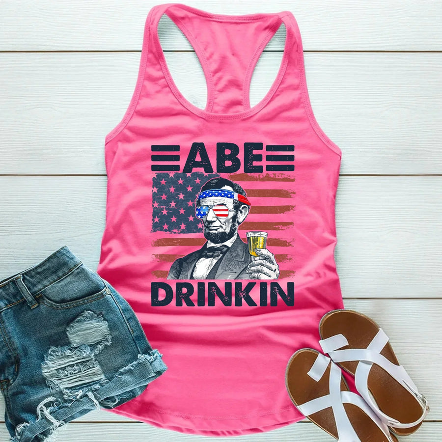Pink tank top with Abraham Lincoln graphic wearing American flag sunglasses, holding a drink, paired with denim shorts and sandals.