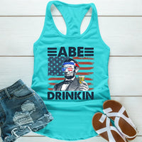 Turquoise tank top with Abraham Lincoln graphic, USA flag, and "Abe Drinkin" text, laid with denim shorts and sandals.