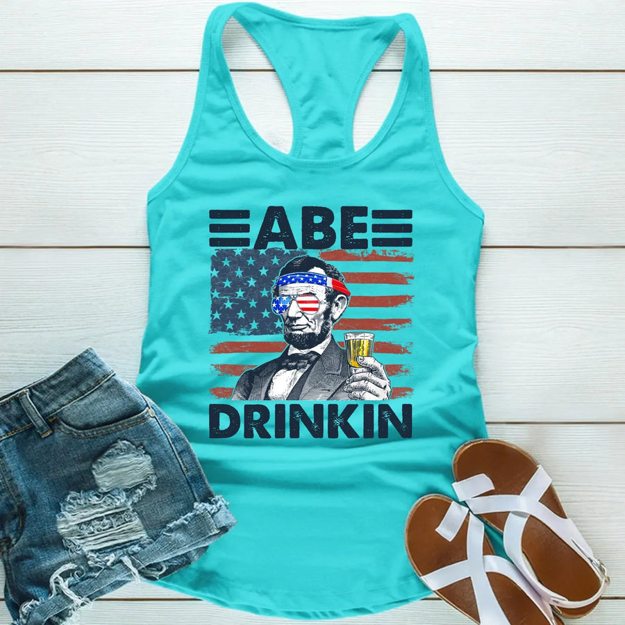 Turquoise tank top with Abraham Lincoln graphic, USA flag, and "Abe Drinkin" text, laid with denim shorts and sandals.