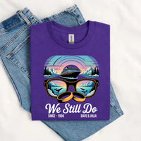 Purple custom tee with cruise ship, sunglasses, wedding rings, and 'We Still Do' text, laid on jeans