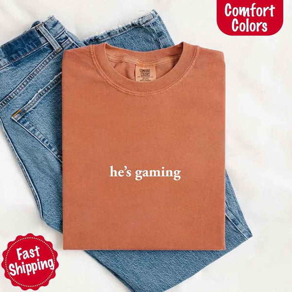 May include a folded yam pastel Comfort Colors T-shirt with the phrase “he’s grilling” printed in black, placed on denim jeans for a clean, casual presentation.