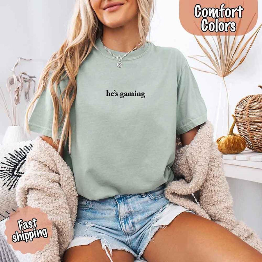May include a woman wearing yam a pastel Comfort Colors T-shirt with the phrase “he’s grilling” printed in black text, styled casually for a fun summer look.