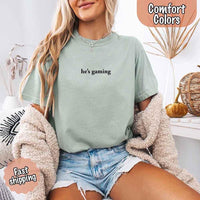 May include a woman wearing yam a pastel Comfort Colors T-shirt with the phrase “he’s grilling” printed in black text, styled casually for a fun summer look.
