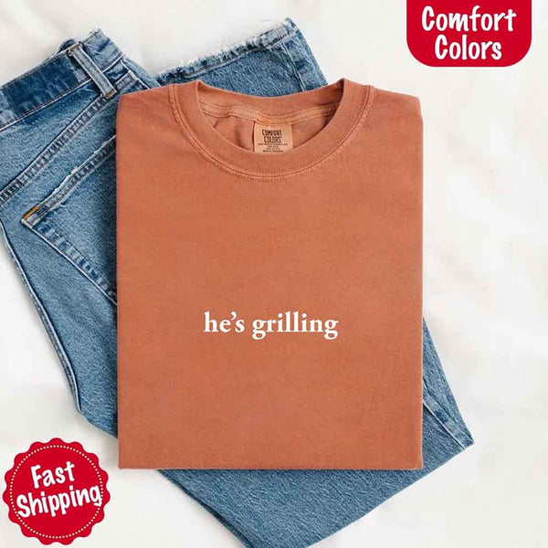 May include a folded yam Comfort Colors T-shirt with “he’s grilling” printed in black, laid over denim jeans for a relaxed summer look.