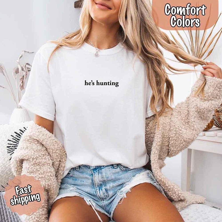May include a woman wearing a white Comfort Colors T-shirt with the phrase “he’s hunting” in black text, styled casually for an outdoor or country-inspired look.