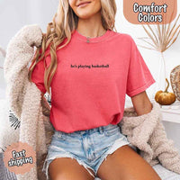 May include a woman wearing a crunchberry Comfort Colors T-shirt with the phrase “he’s playing basketball” in black text, styled casually for a sporty and fun girlfriend look.
