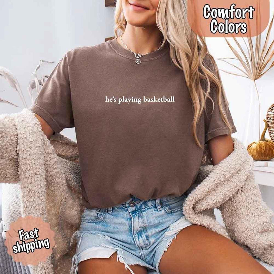 Person wearing a brown t-shirt with text and denim shorts, sitting on a couch wearing a espresso comfort colors t-shirt with 'he's playing basketball' text on blue jeans, with 'Comfort Colors' and 'Fast Shipping' logos.