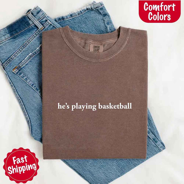Folded brown t-shirt with 'he's playing basketball' text on blue jeans, with 'Comfort Colors' and 'Fast Shipping' logos.
