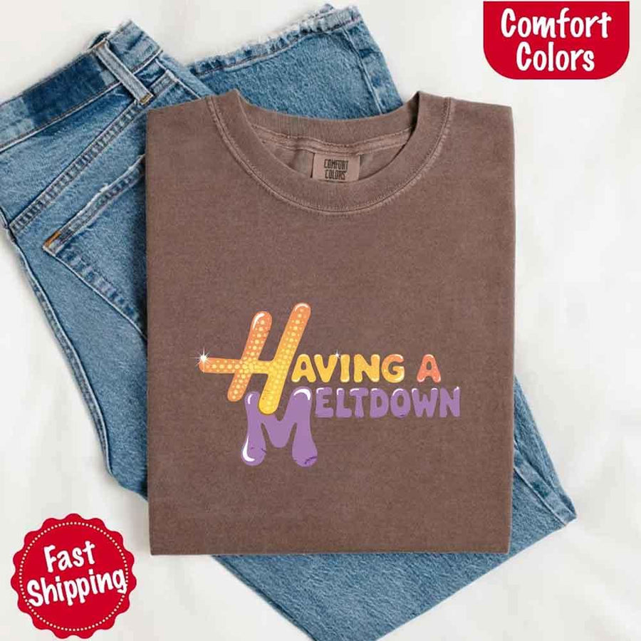 May include a folded espresso Comfort Colors T-shirt with “having a meltdown” printed in black, placed over denim jeans for a playful and relaxed vibe.