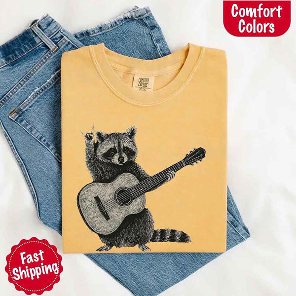 May include a folded mustard Comfort Colors T-shirt with a cute raccoon graphic holding a guitar, laid over denim jeans for a playful and humorous vibe.