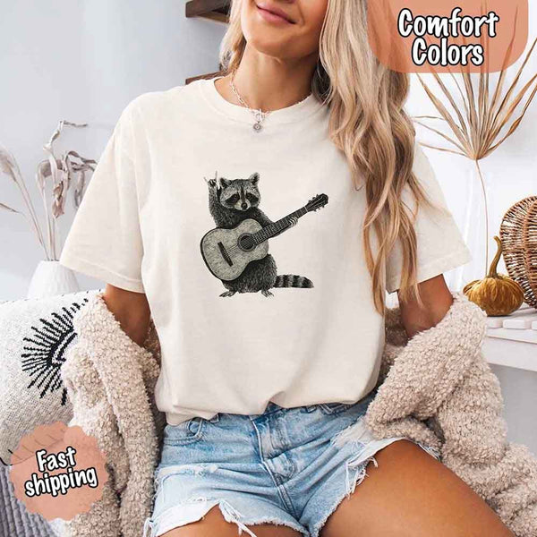 May include a woman wearing a ivory Comfort Colors T-shirt featuring a graphic of a raccoon playing a guitar, styled for a quirky and fun animal lover look.